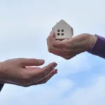 Mão entregando uma pequena casa de modelo para outra pessoa, simbolizando doação ou transferência de propriedade.