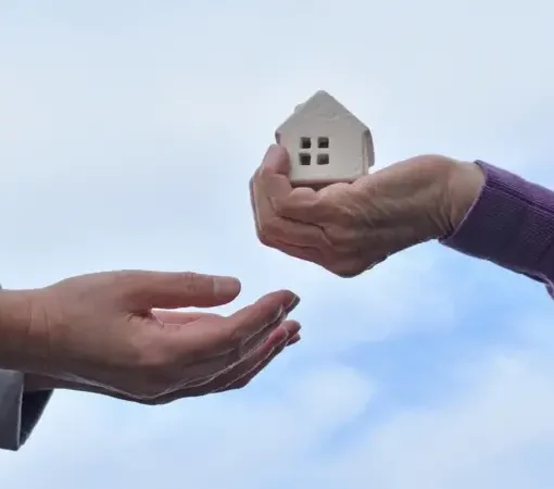 Mão entregando uma pequena casa de modelo para outra pessoa, simbolizando doação ou transferência de propriedade.