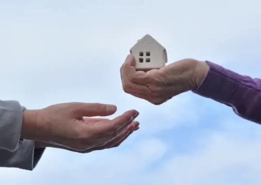 Mão entregando uma pequena casa de modelo para outra pessoa, simbolizando doação ou transferência de propriedade.